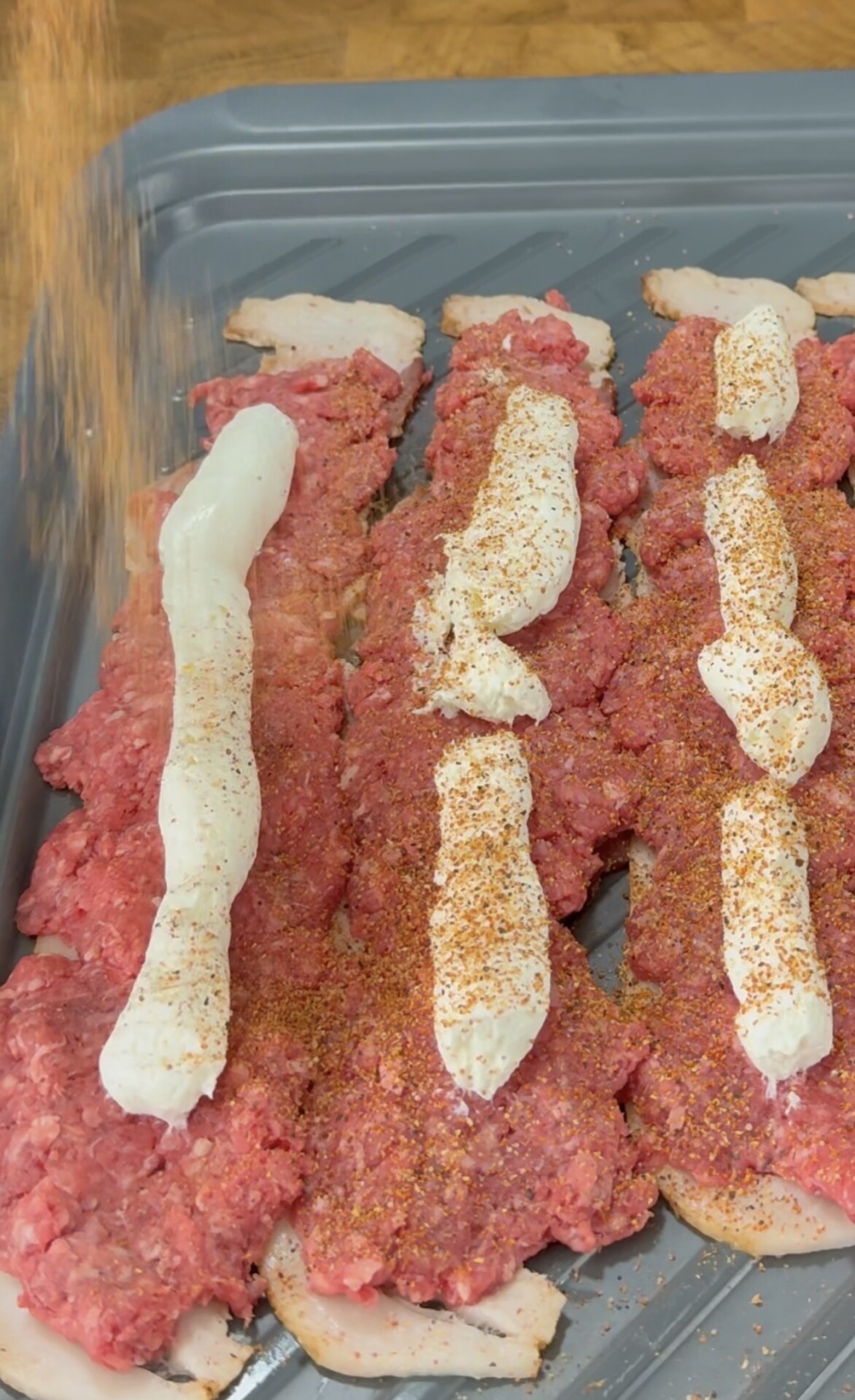 Rows of raw ground meat are lined up on a baking tray, each topped with strips of white creamy sauce and being sprinkled with a reddish seasoning.
