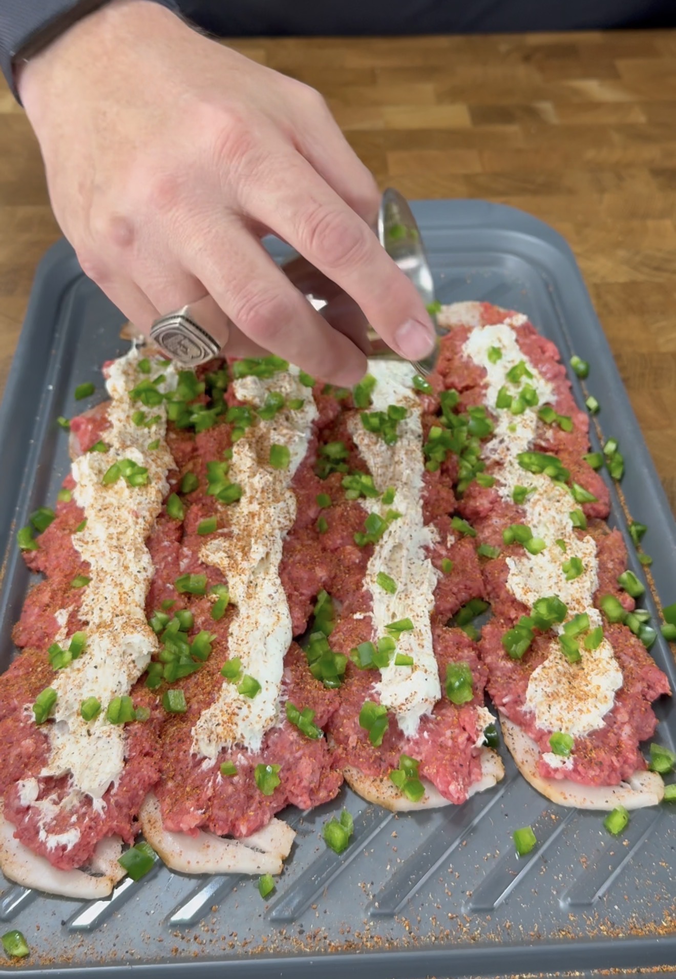 A hand sprinkles chopped green peppers onto rows of raw ground meat mixture topped with a white creamy sauce on a baking tray, preparing the dish for cooking.