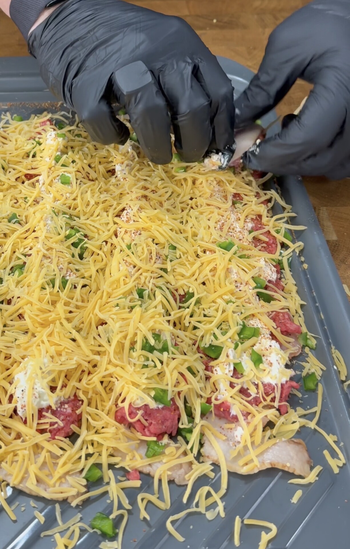 Gloved hands preparing a baking tray filled with raw chicken breasts topped with shredded cheddar cheese, ground meat, diced green peppers, and seasonings.