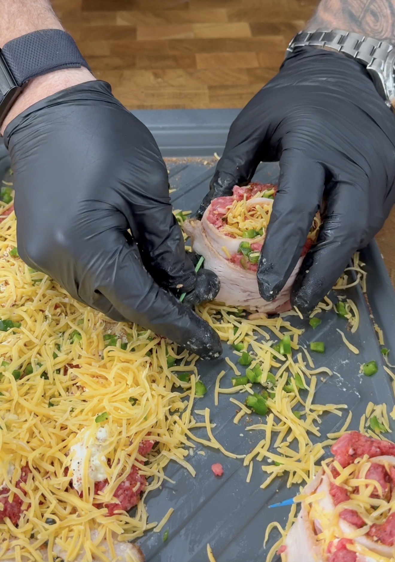 A person wearing black gloves is shaping a bacon-wrapped meat roll topped with shredded cheese and chopped green peppers on a baking tray, with more cheese scattered around.