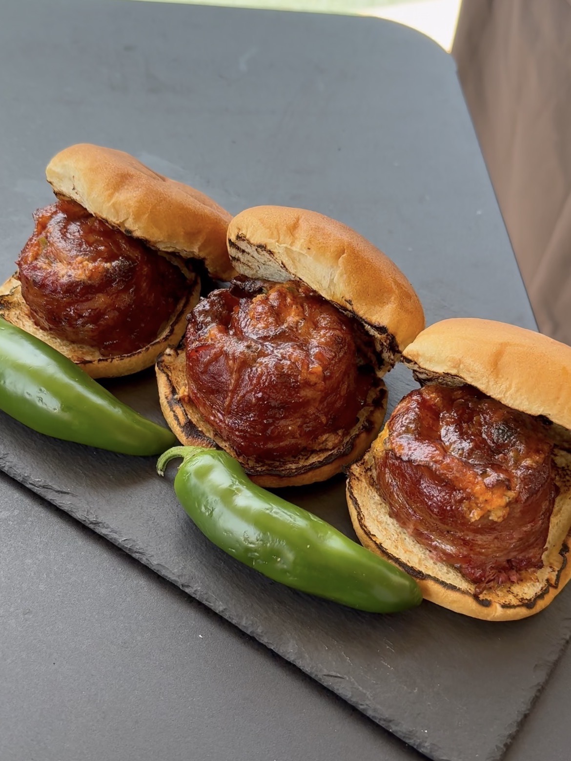Three burgers with thick, bacon-wrapped patties are served on toasted buns, displayed on a black slate, with two whole green jalapeño peppers placed in front of them.