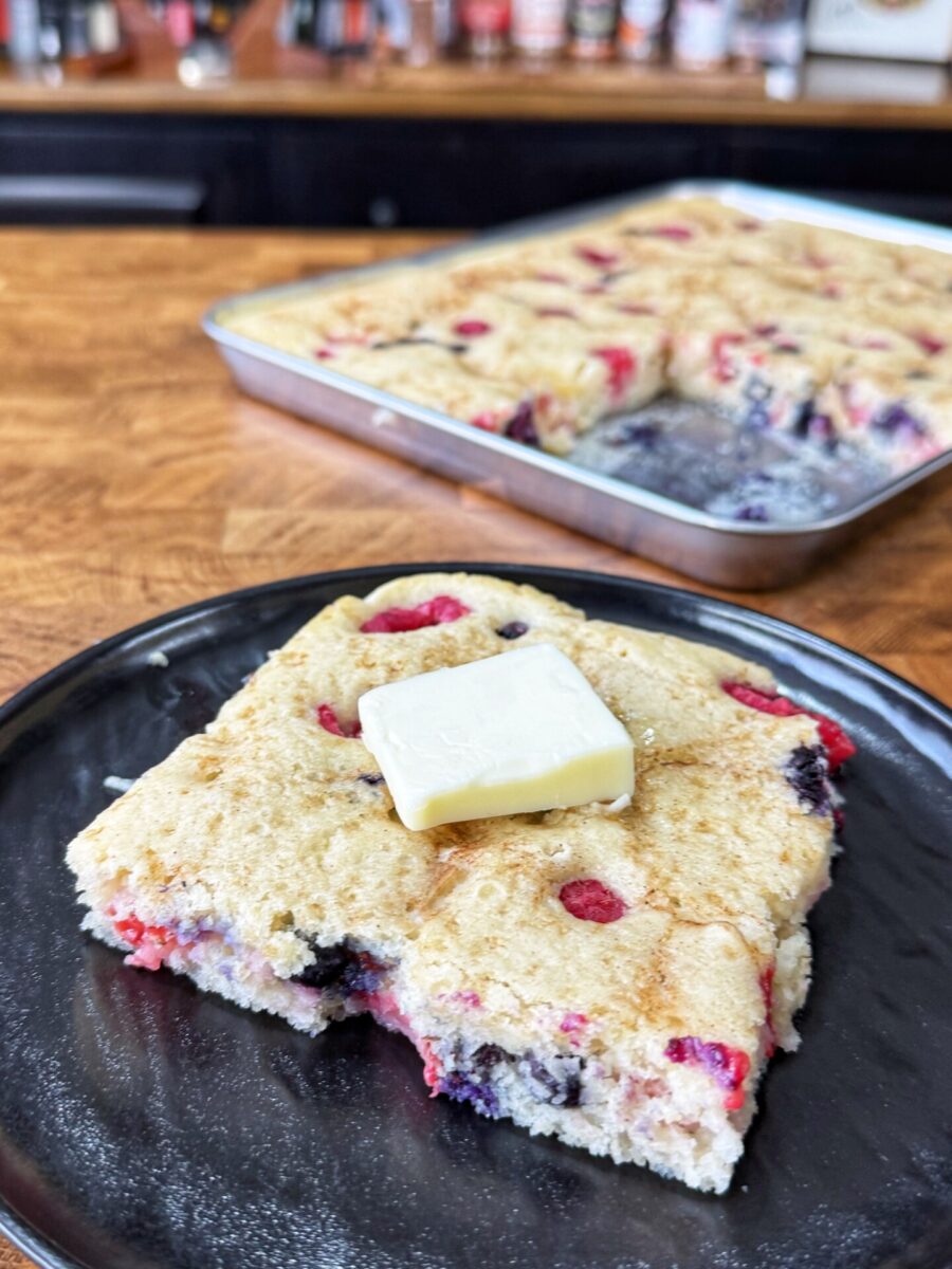 A square piece of berry-studded pancake topped with a pat of butter sits on a black plate, with the remaining pancake in a baking pan in the background on a wooden countertop.