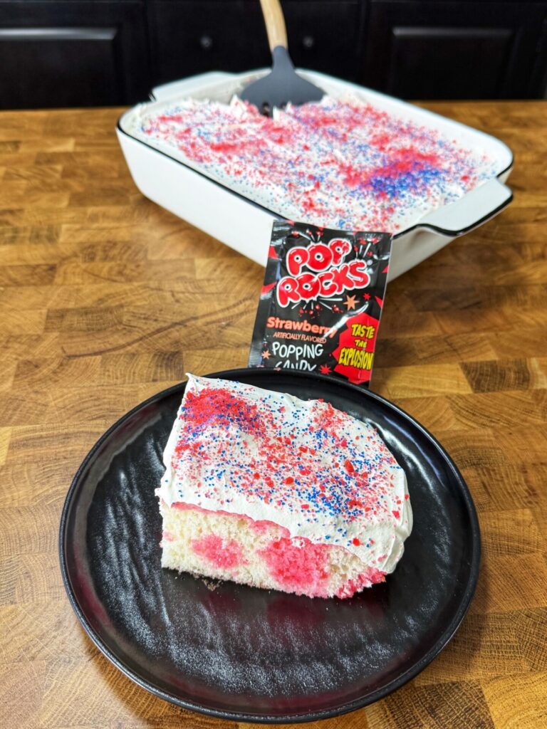 A slice of white cake with pink spots and white frosting, topped with red and blue Pop Rocks candy, sits on a black plate. The rest of the cake is in a white baking dish in the background with a Pop Rocks packet nearby.