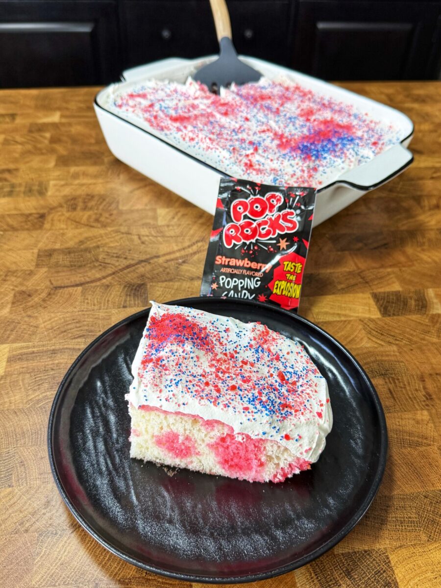 A slice of white cake with pink spots and white frosting, topped with red and blue Pop Rocks candy, sits on a black plate. The rest of the cake is in a white baking dish in the background with a Pop Rocks packet nearby.