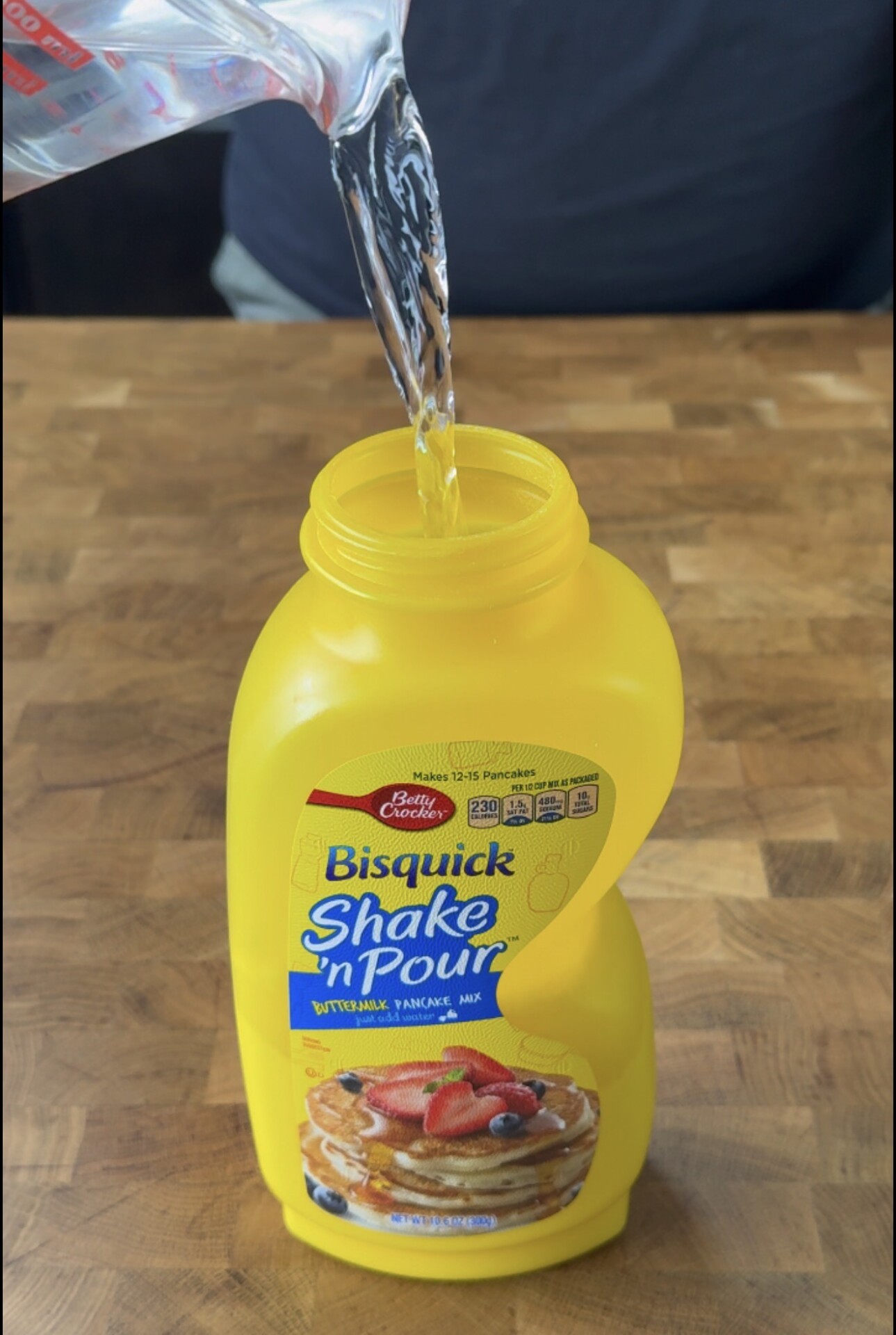 A clear liquid is being poured from a measuring cup into a yellow Bisquick Shake n Pour pancake mix container on a wooden surface.