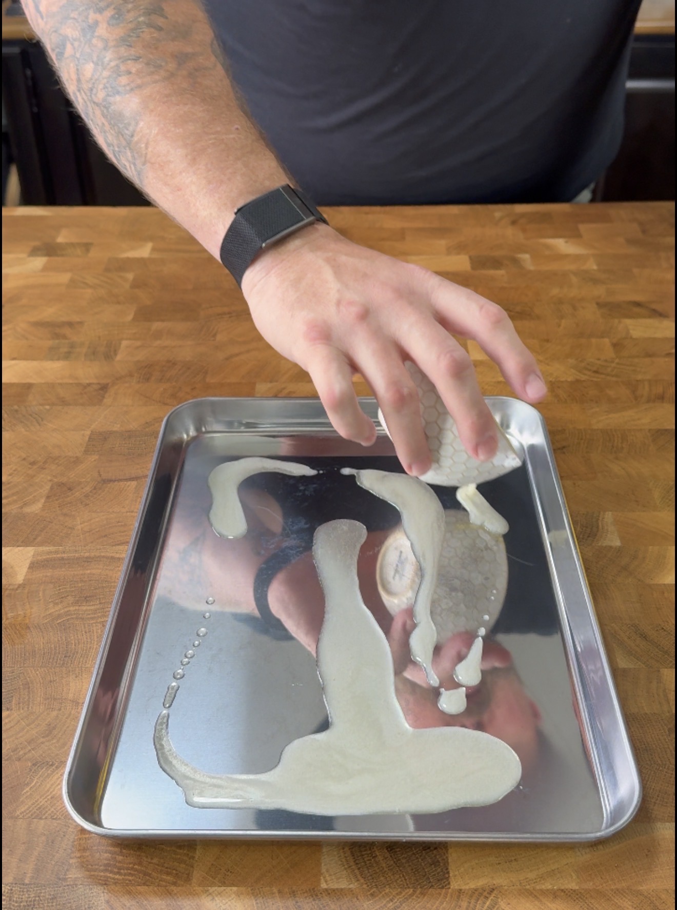 A person wearing a smartwatch is lifting a piece of honeycomb-patterned tripe from a metal tray with a light-colored sauce or puree spread across the tray’s surface. The tray is on a wooden countertop.