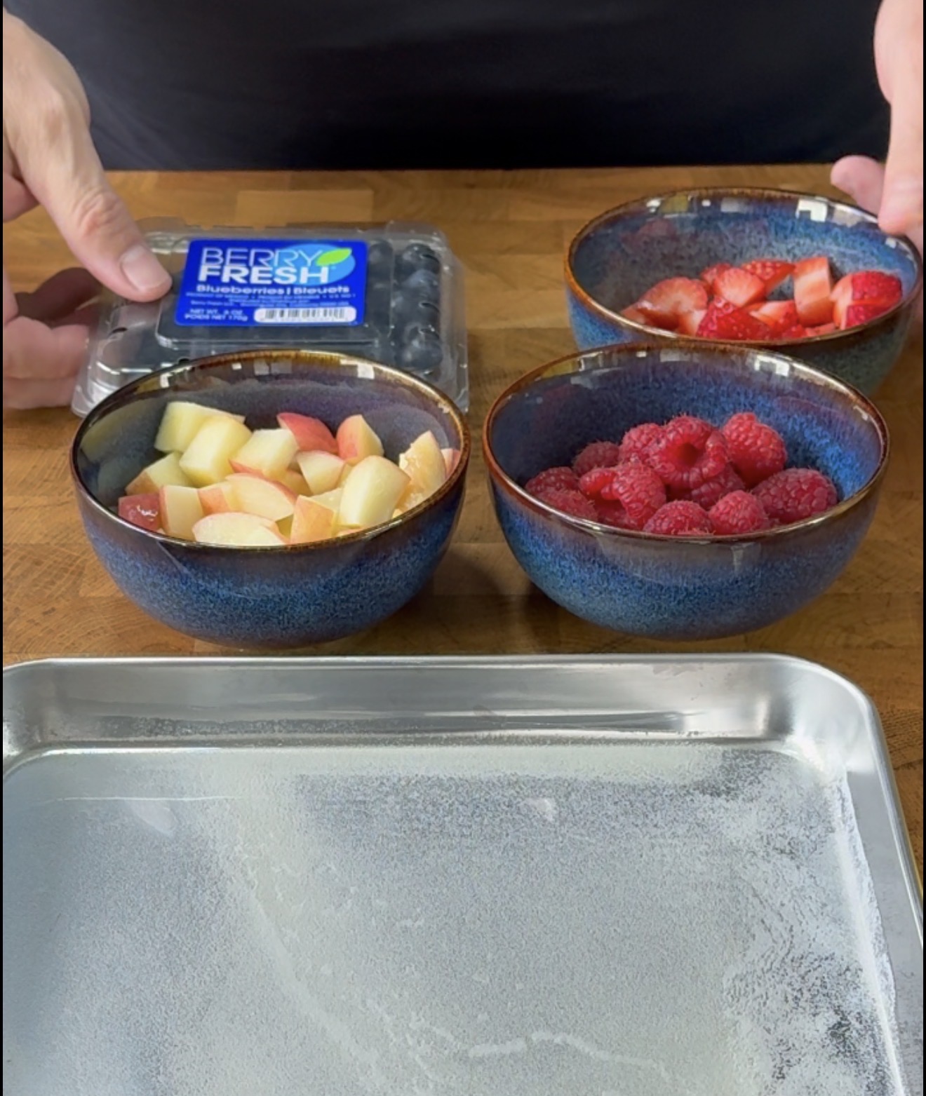A person arranges bowls of chopped apples, raspberries, and strawberries beside a container of blueberries on a wooden counter, with a metal tray lined with parchment paper in front.