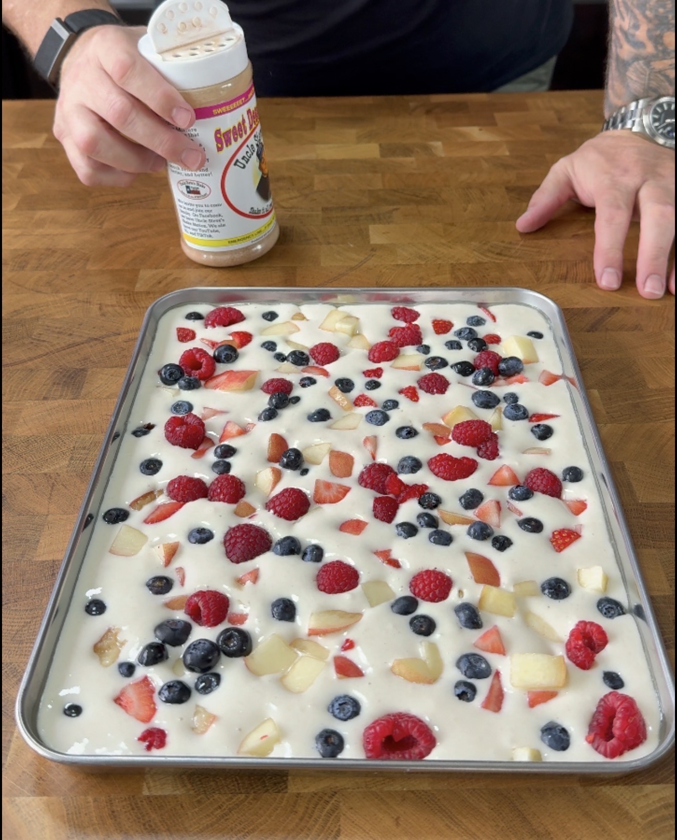 A person sprinkles seasoning onto a baking tray filled with uncooked batter topped with raspberries, blueberries, strawberries, and apple pieces on a wooden surface.