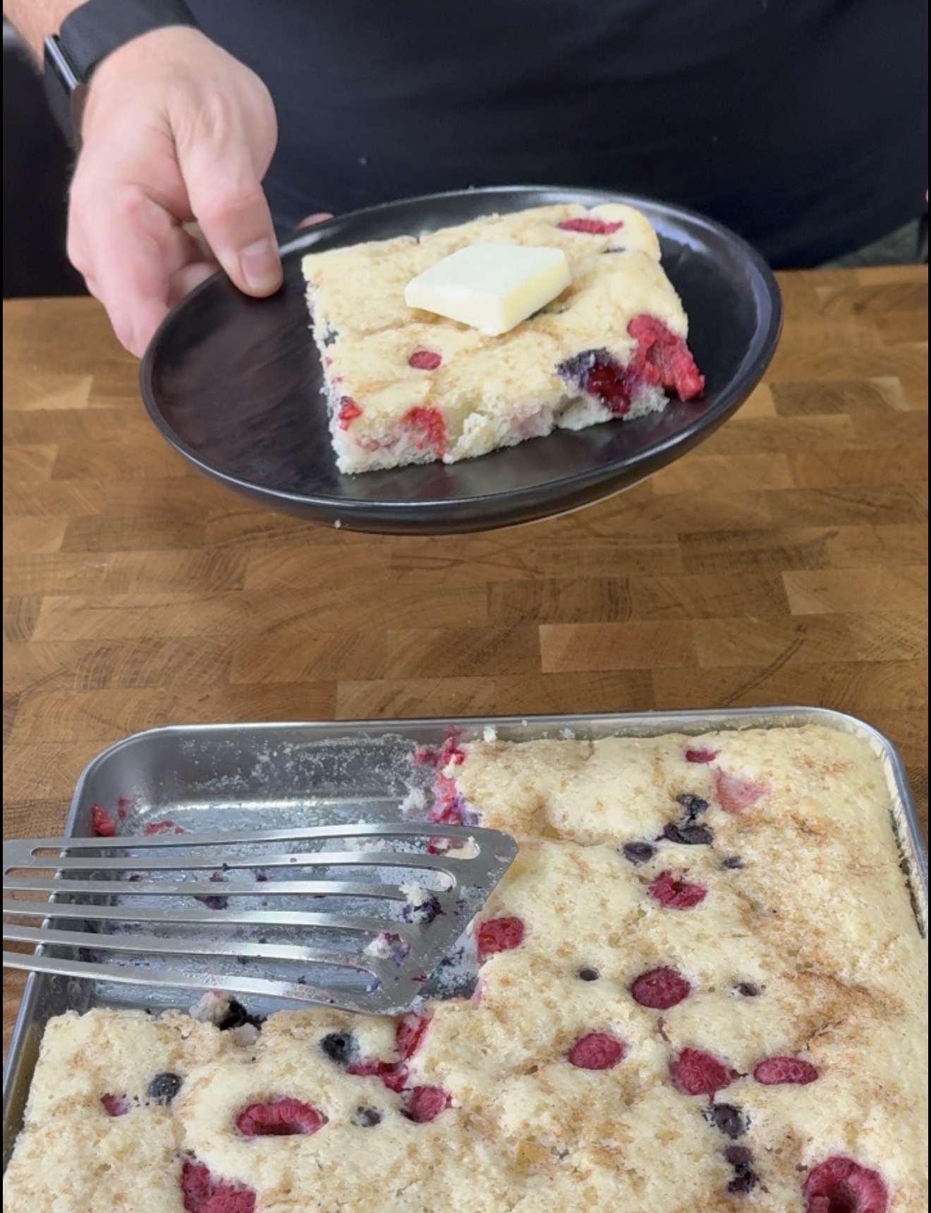 A person holds a plate with a square piece of berry-studded baked pancake topped with a pat of butter; the rest of the pancake is in a pan below with a spatula resting on it.