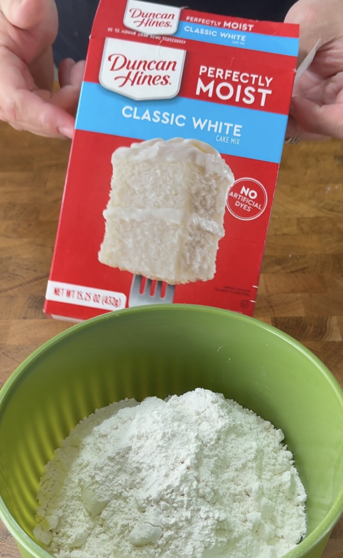 A person holds a box of Duncan Hines Classic White cake mix above a green bowl filled with the dry cake mix on a wooden surface.