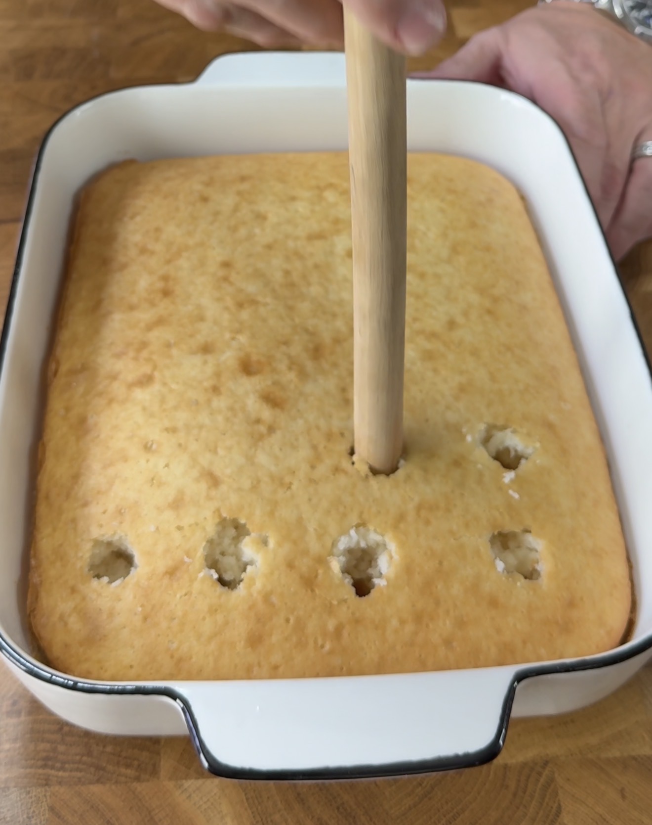 A hand uses the handle of a wooden spoon to poke holes in a rectangular baked cake in a white baking dish on a wooden countertop.