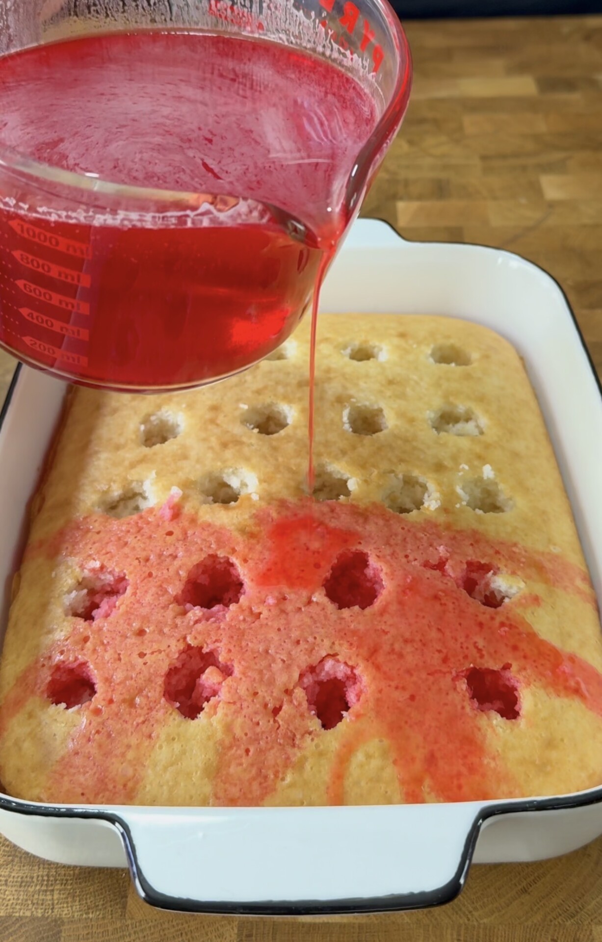 A close-up of red liquid being poured from a measuring cup onto a yellow poke cake with holes in a white baking dish, set on a wooden surface.