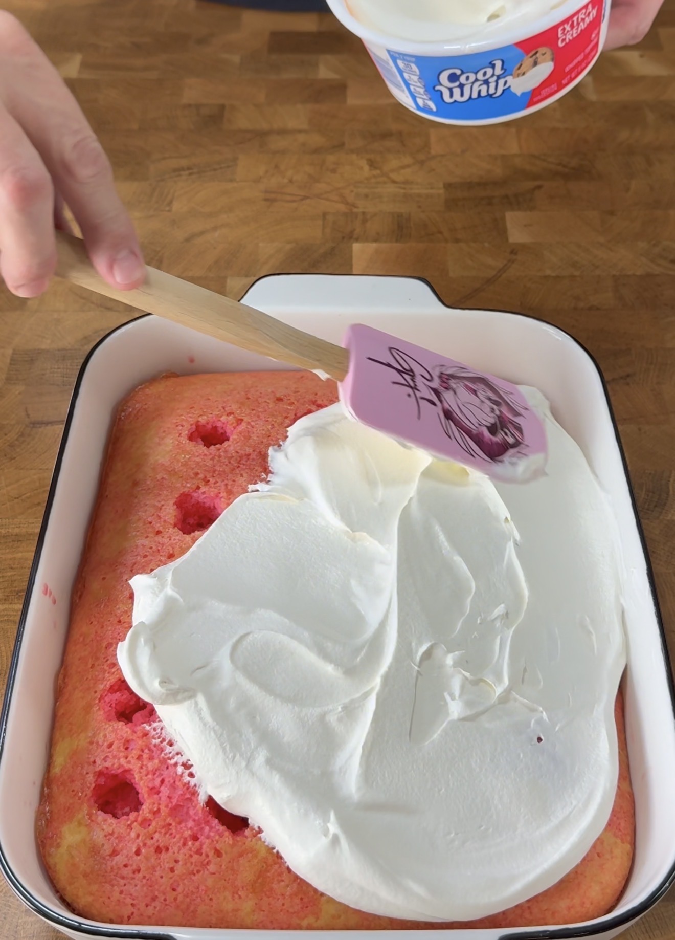 A hand uses a pink spatula to spread white whipped topping over a rectangular pink cake with holes, in a white baking dish on a wooden surface. A tub of Cool Whip is in the background.