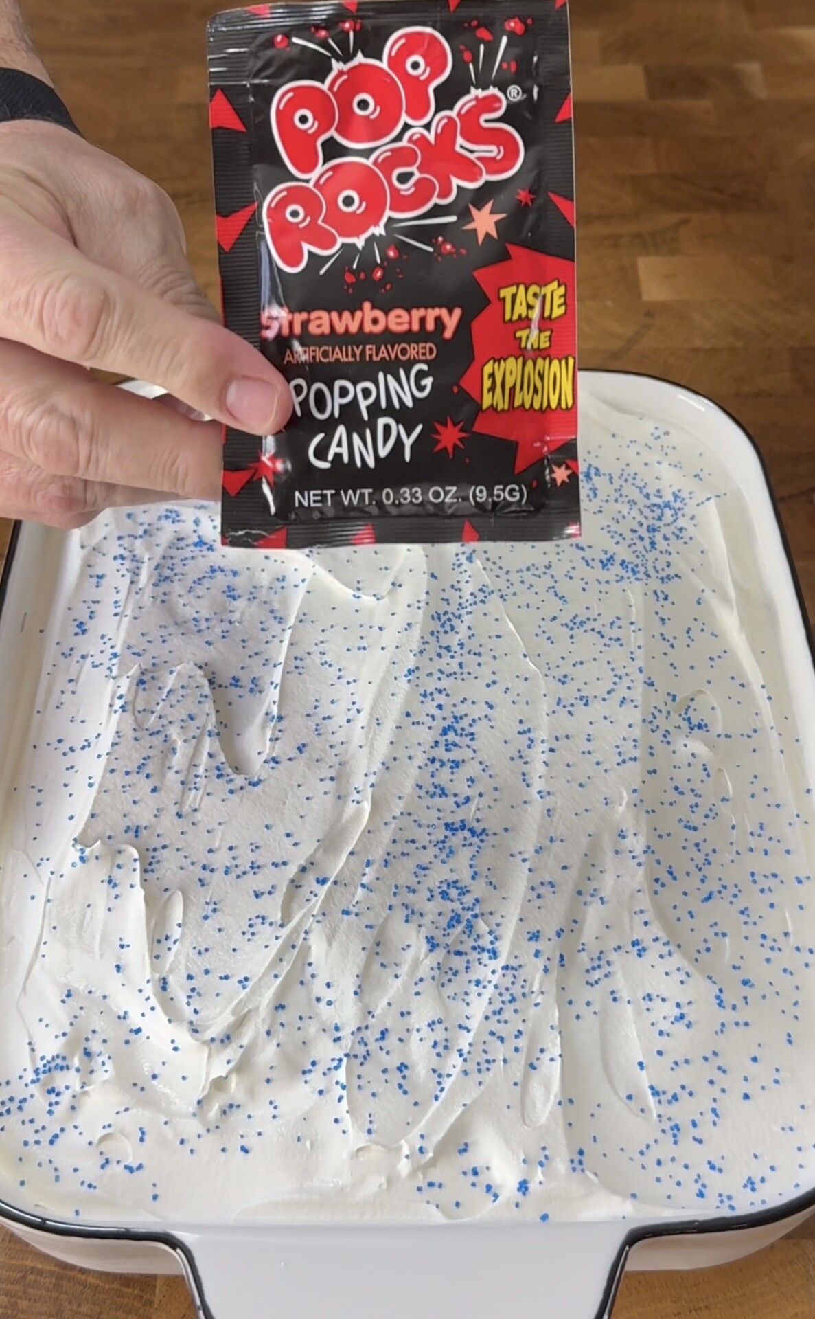 A hand holds a packet of strawberry Pop Rocks above a dish of dessert topped with white frosting and blue sprinkles on a wooden surface.