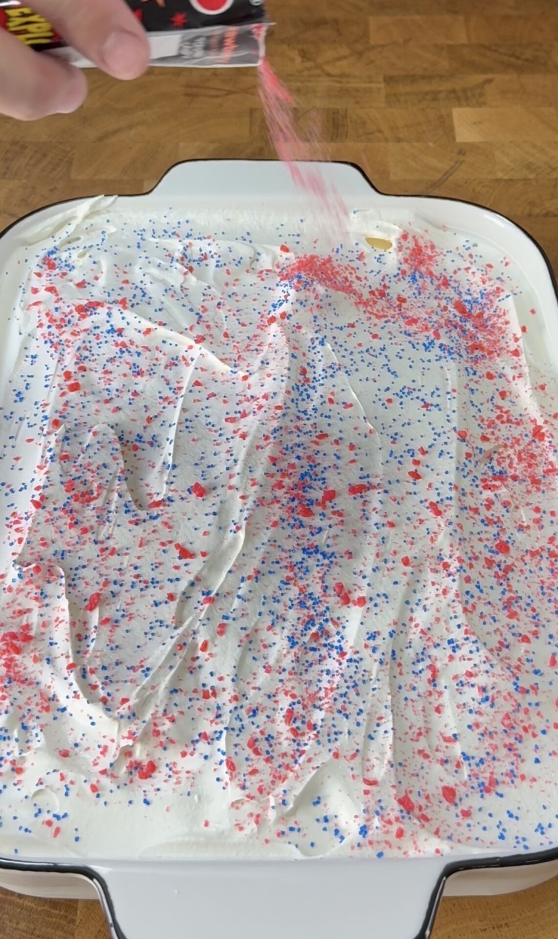A hand sprinkles red and blue Pop Rocks candy over a rectangular dish covered with a layer of white whipped topping on a wooden surface.