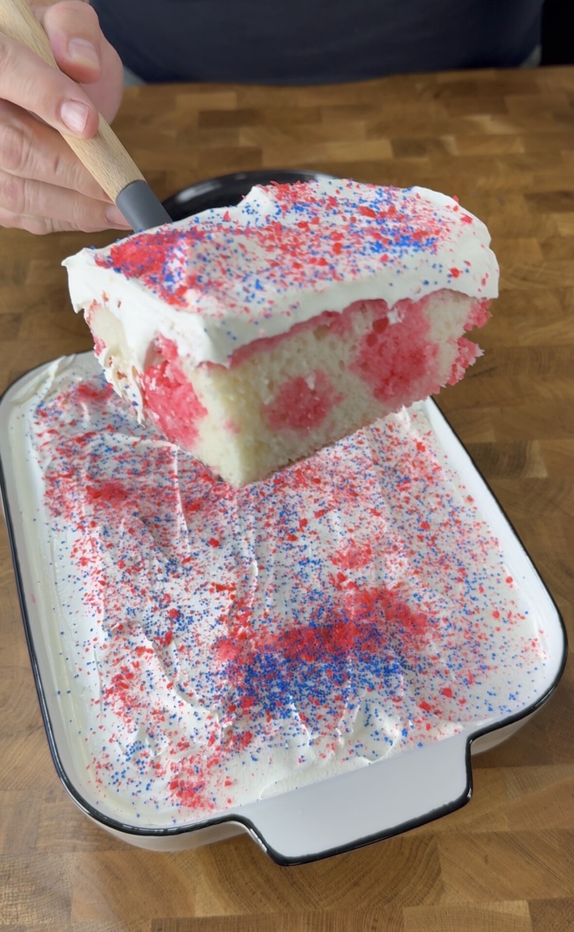 A slice of white poke cake with red gelatin spots, topped with white whipped cream and red and blue sprinkles, is lifted from a rectangular dish on a wooden surface.
