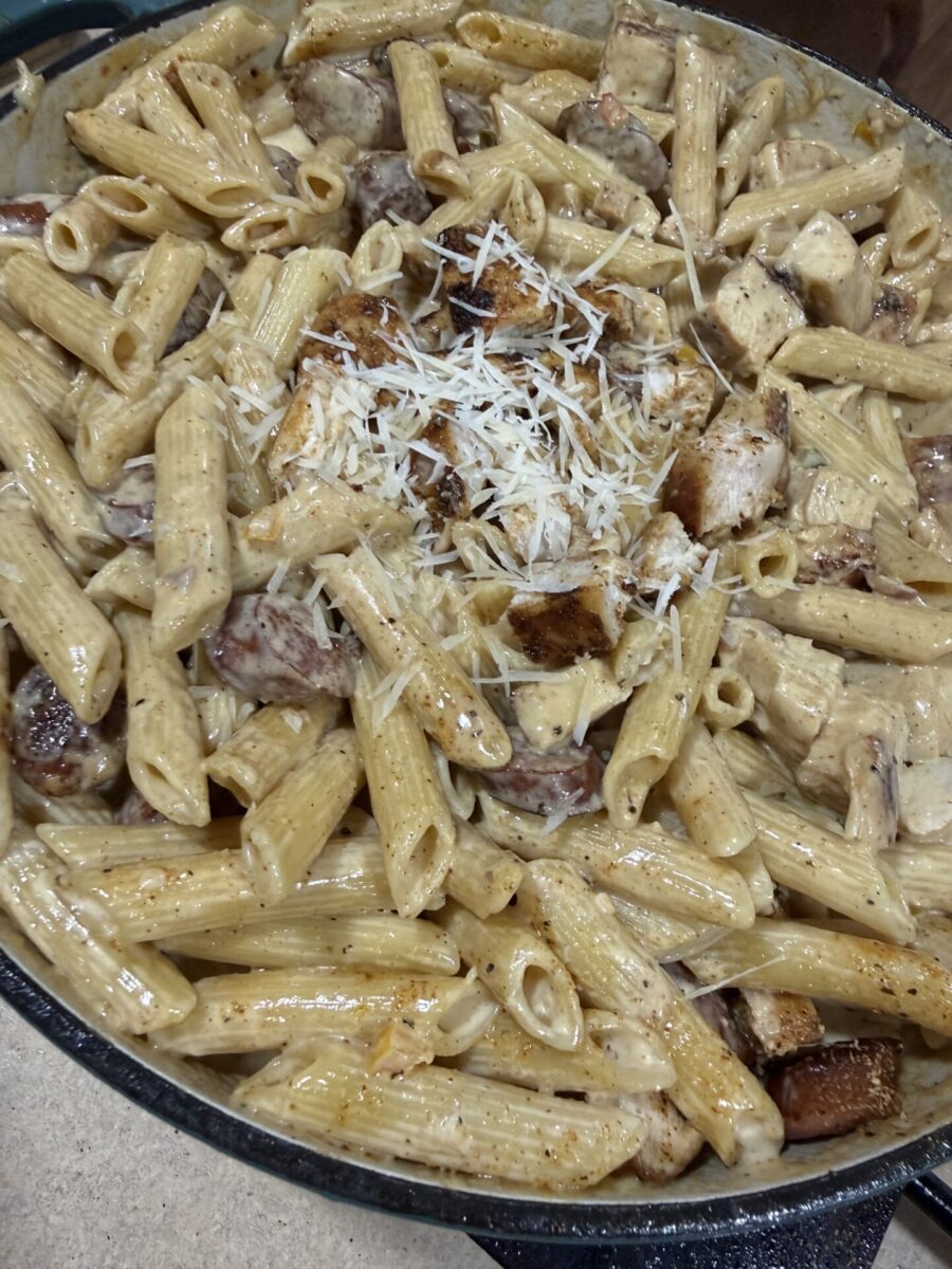A close-up of creamy penne pasta mixed with grilled chicken and sausage slices, topped with shredded parmesan cheese, served in a large pan.