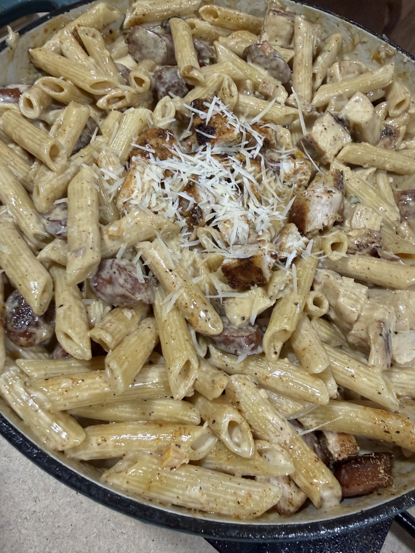 A close-up of creamy penne pasta mixed with grilled chicken and sausage slices, topped with shredded parmesan cheese, served in a large pan.