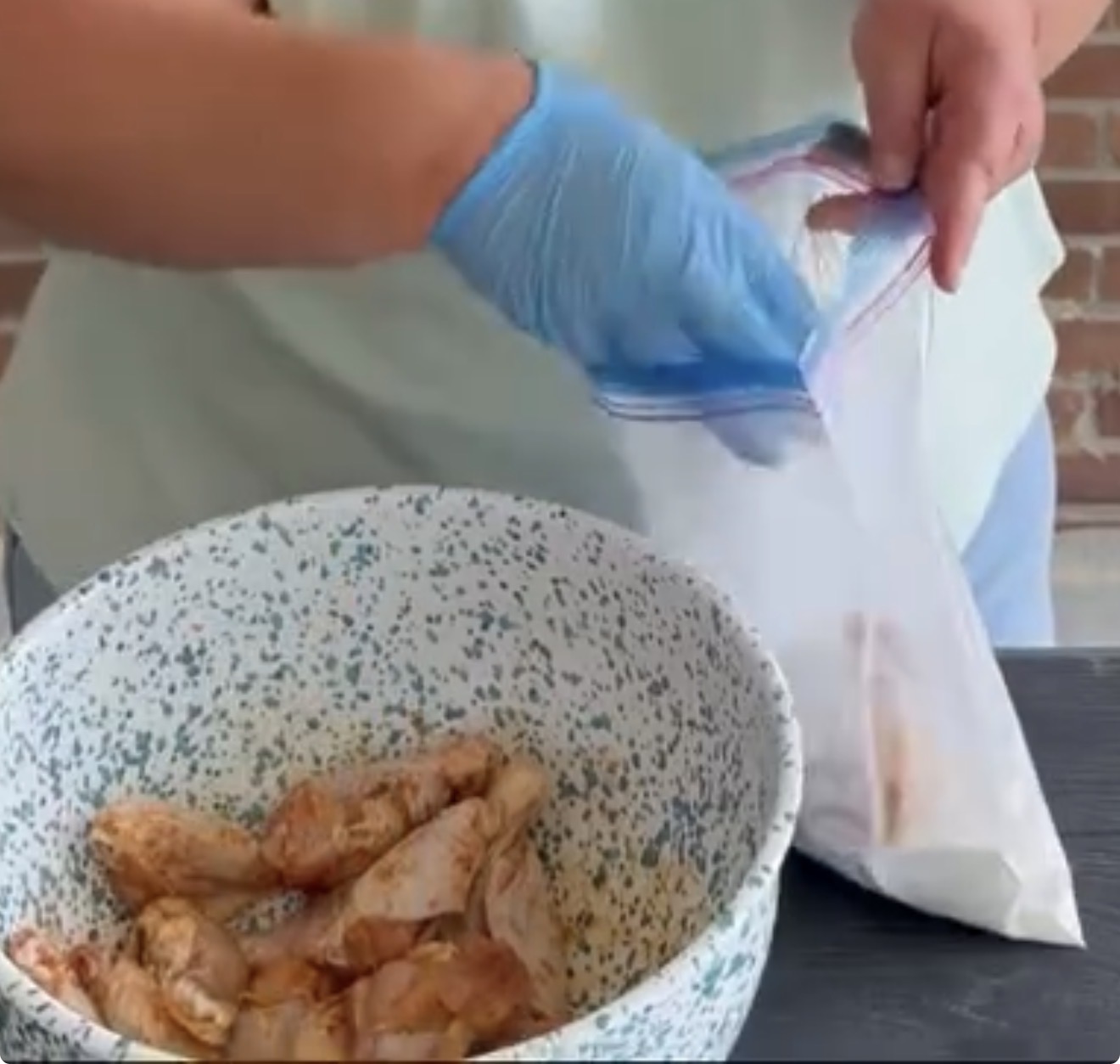A person wearing a blue glove places seasoned chicken drumsticks into a clear ziplock bag above a speckled mixing bowl containing more chicken pieces.