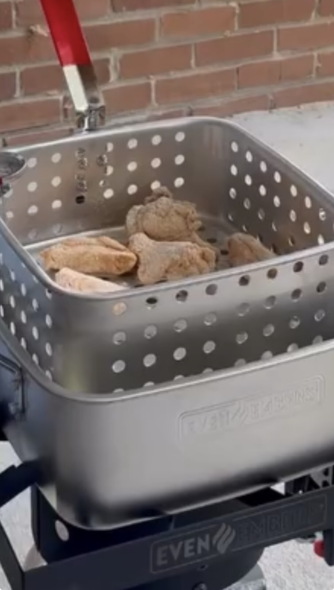 A metal basket containing several pieces of seasoned raw chicken sits on an outdoor deep fryer, with a brick wall in the background.