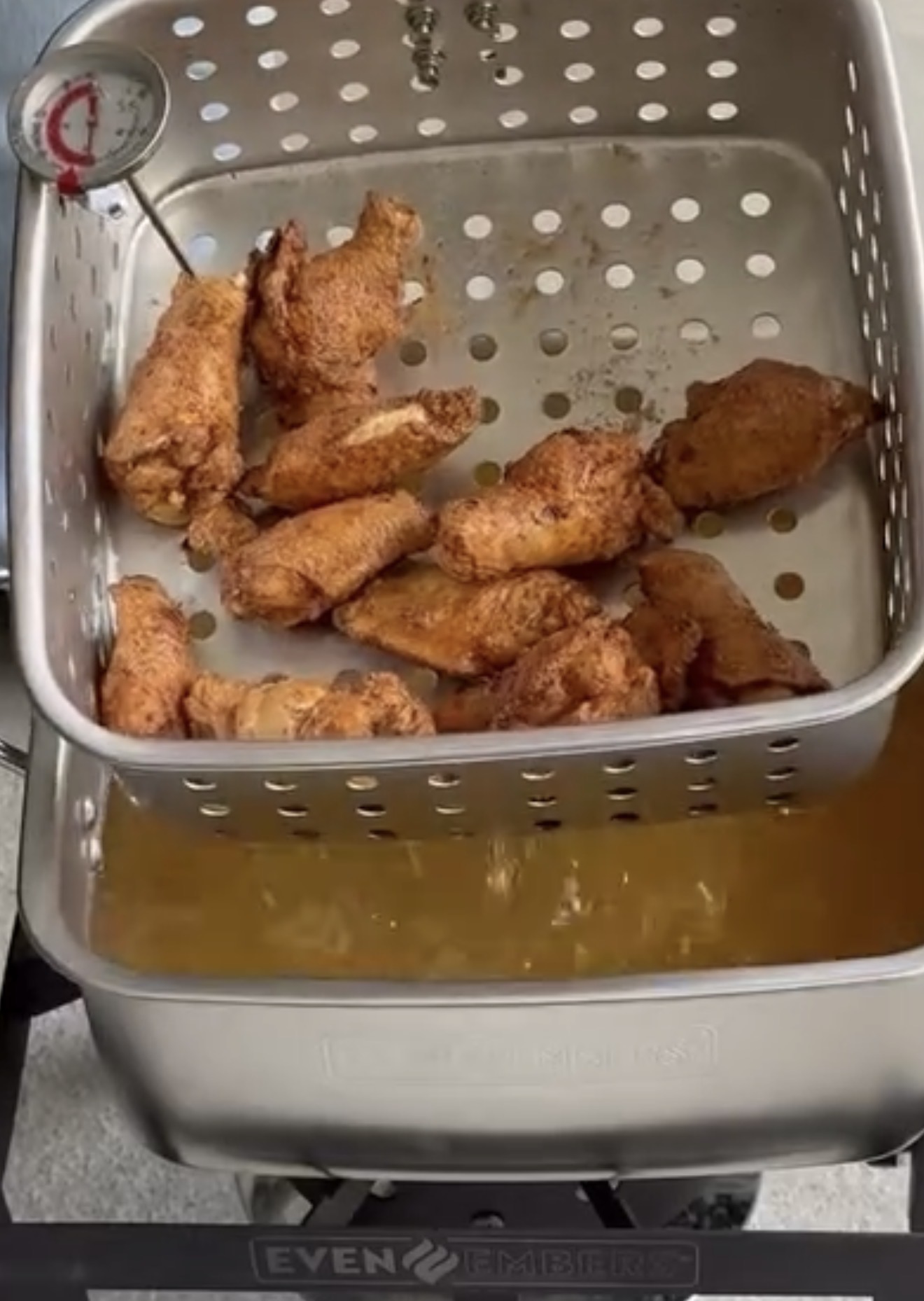 A metal basket filled with fried chicken wings is being lifted out of a deep fryer, with a thermometer inserted in the food and oil visible below the basket.