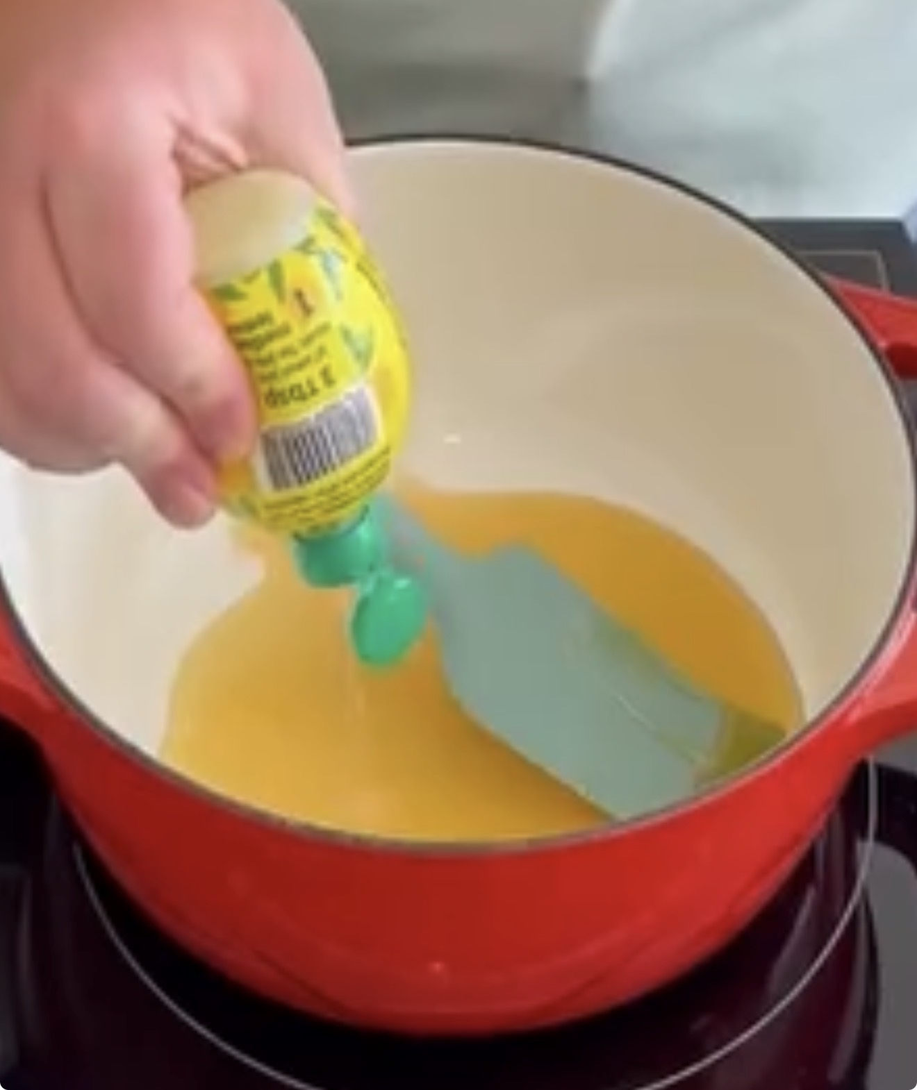 A hand squeezes lemon juice from a plastic bottle into a red pot on a stovetop, with a blue spatula stirring the yellow liquid inside.