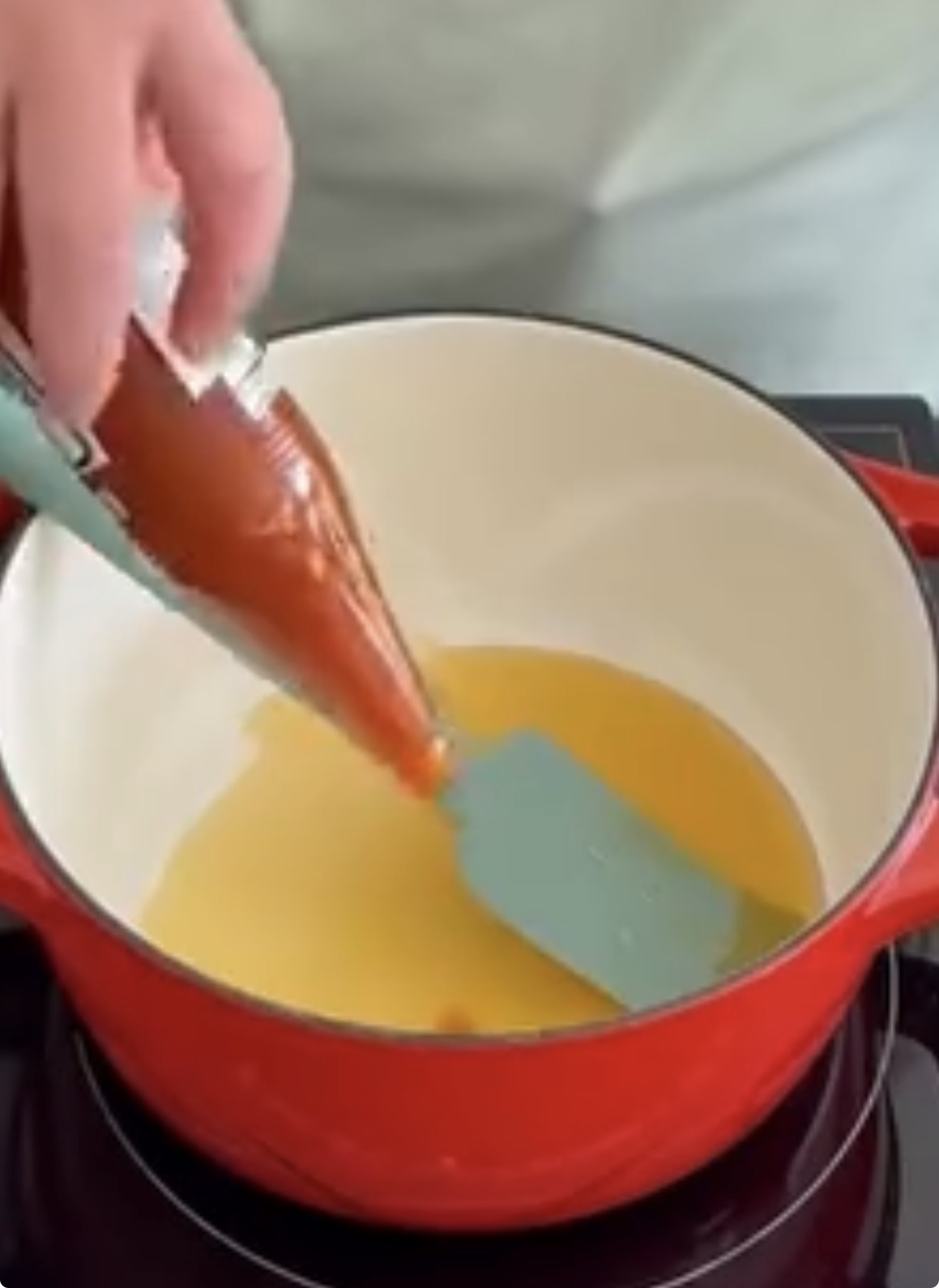 A hand squeezes a bottle of hot sauce into a red pot containing melted butter on a stovetop. A blue spatula is in the pot, ready for stirring.
