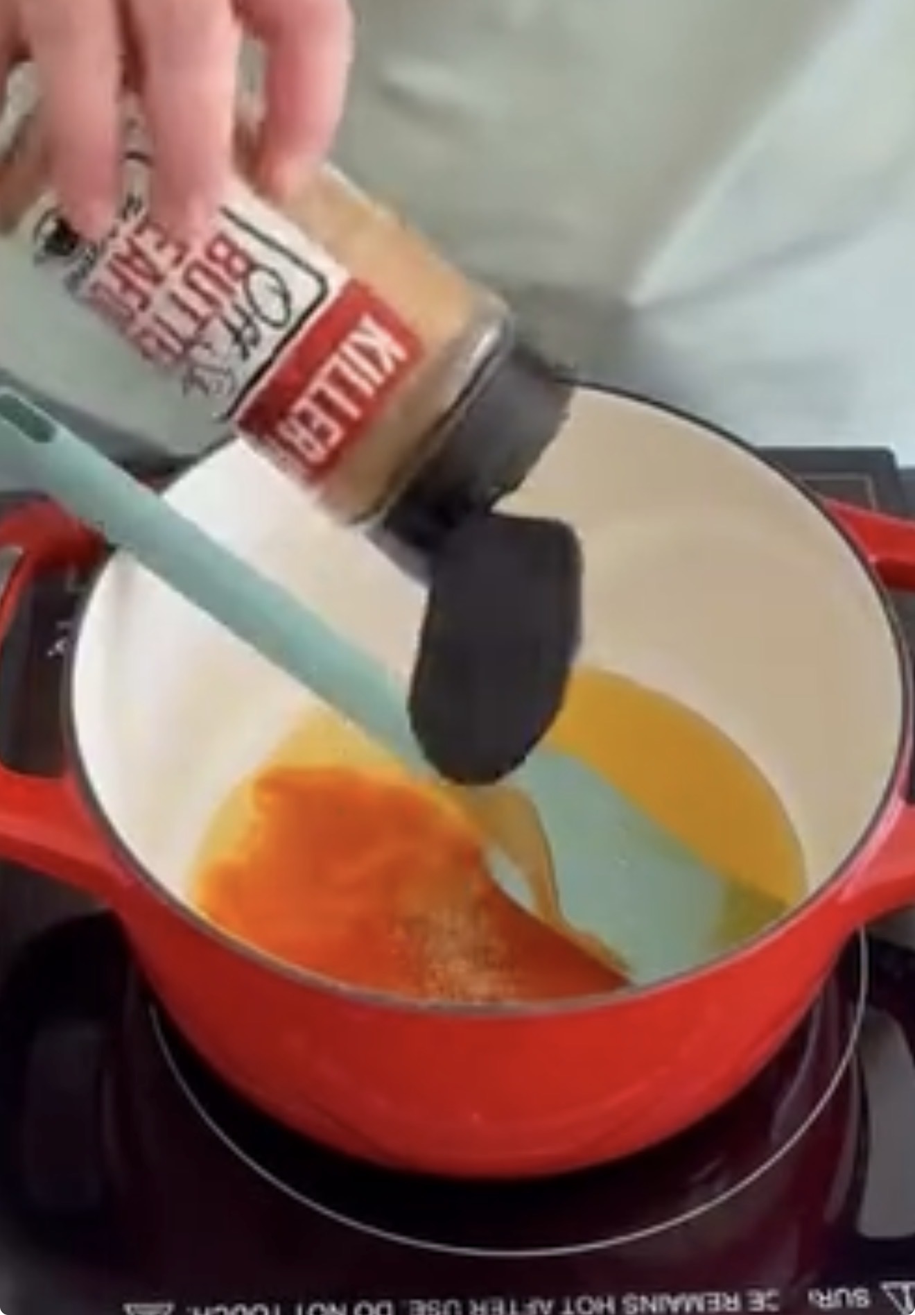 A hand shakes a spice container labeled Killer Bee Honey Rub over a red pot on a stovetop, mixing it with melted butter or oil using a blue spatula.