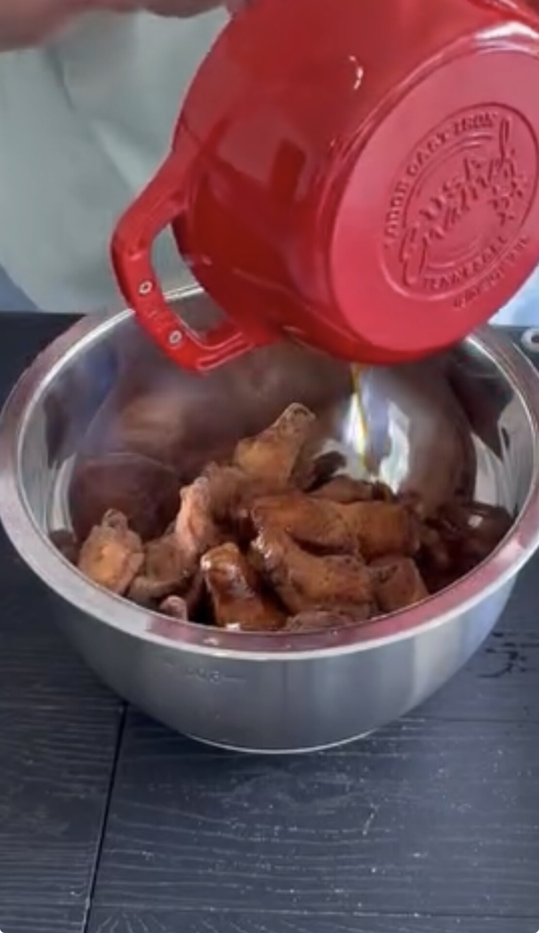 A person pours a dark marinade from a red pot into a metal bowl filled with pieces of raw meat on a dark tabletop.