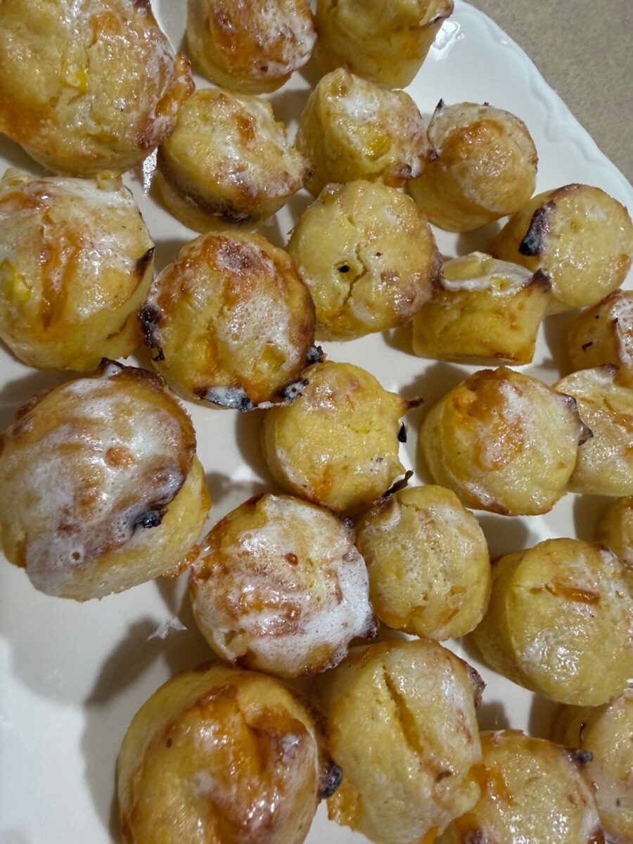 A plate filled with homemade muffins or cupcakes, topped with a light glaze. The baked treats are golden brown with some dark spots, likely from chocolate chips or dried fruit.