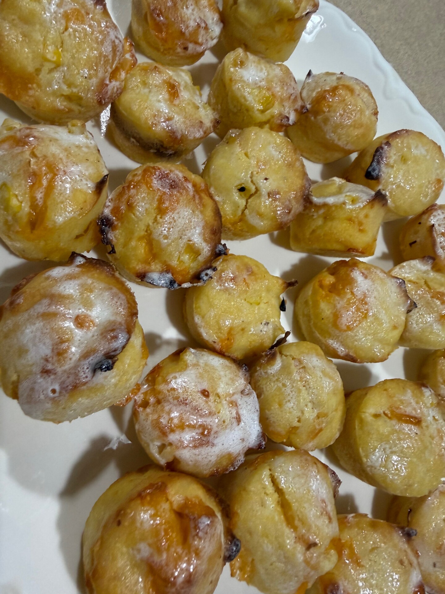 A plate filled with homemade muffins or cupcakes, topped with a light glaze. The baked treats are golden brown with some dark spots, likely from chocolate chips or dried fruit.