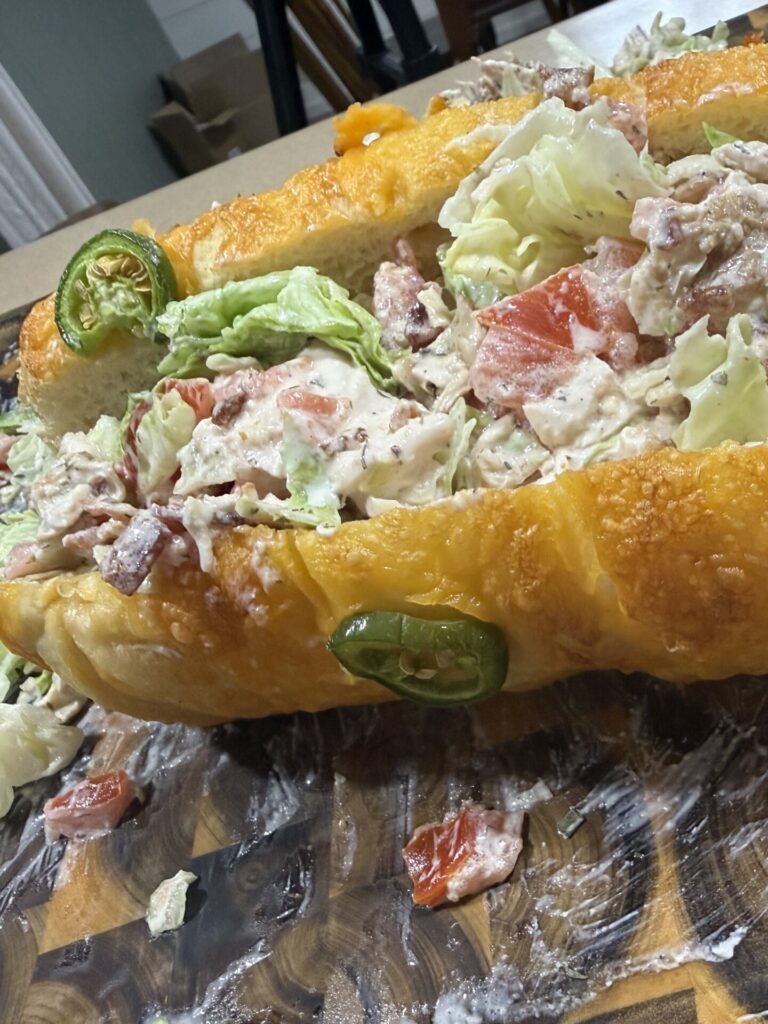 A close-up of a large sandwich on a wooden board, filled with lettuce, tomatoes, bacon, and creamy dressing, topped with sliced jalapeños. Some filling is spilling out onto the board.