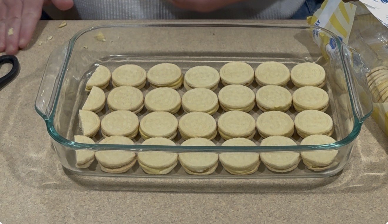 A glass baking dish filled with a single layer of vanilla sandwich cookies arranged neatly in rows on a kitchen countertop. A persons hands and an open package of cookies are visible in the background.