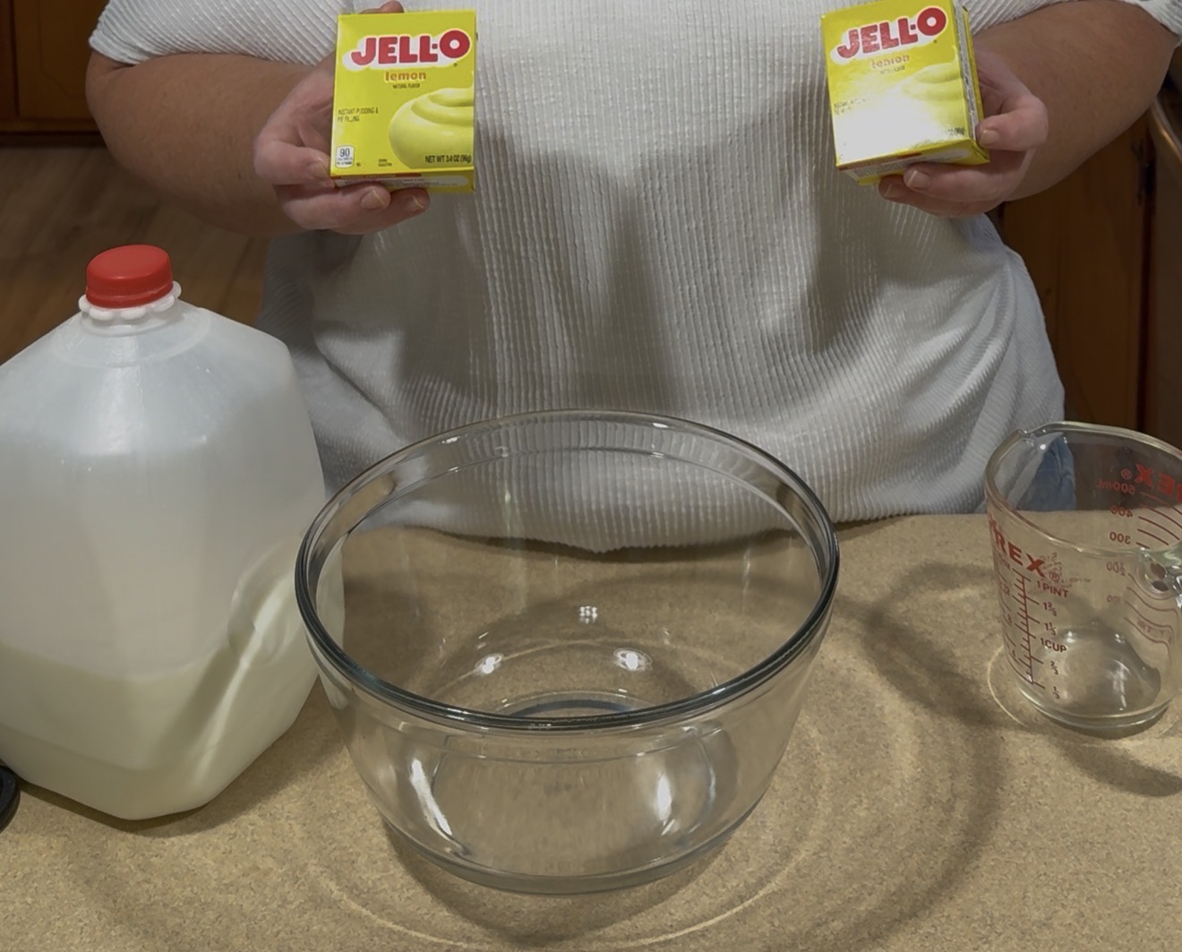 A person holds two boxes of lemon Jello over a clear glass mixing bowl. A gallon of milk and a measuring cup are on the counter nearby.