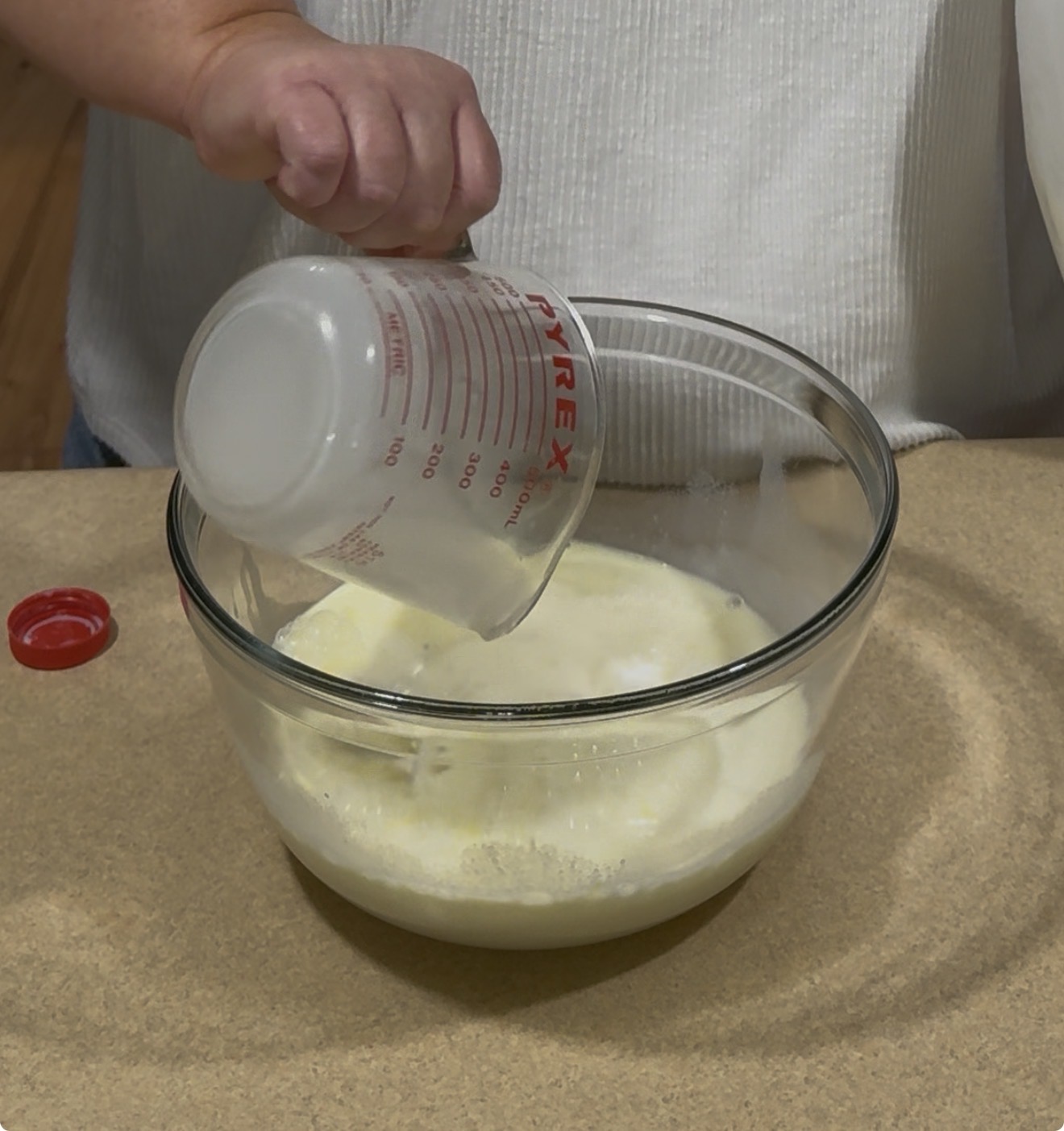 A person pours liquid from a glass measuring cup into a clear glass mixing bowl containing a creamy mixture on a kitchen counter.