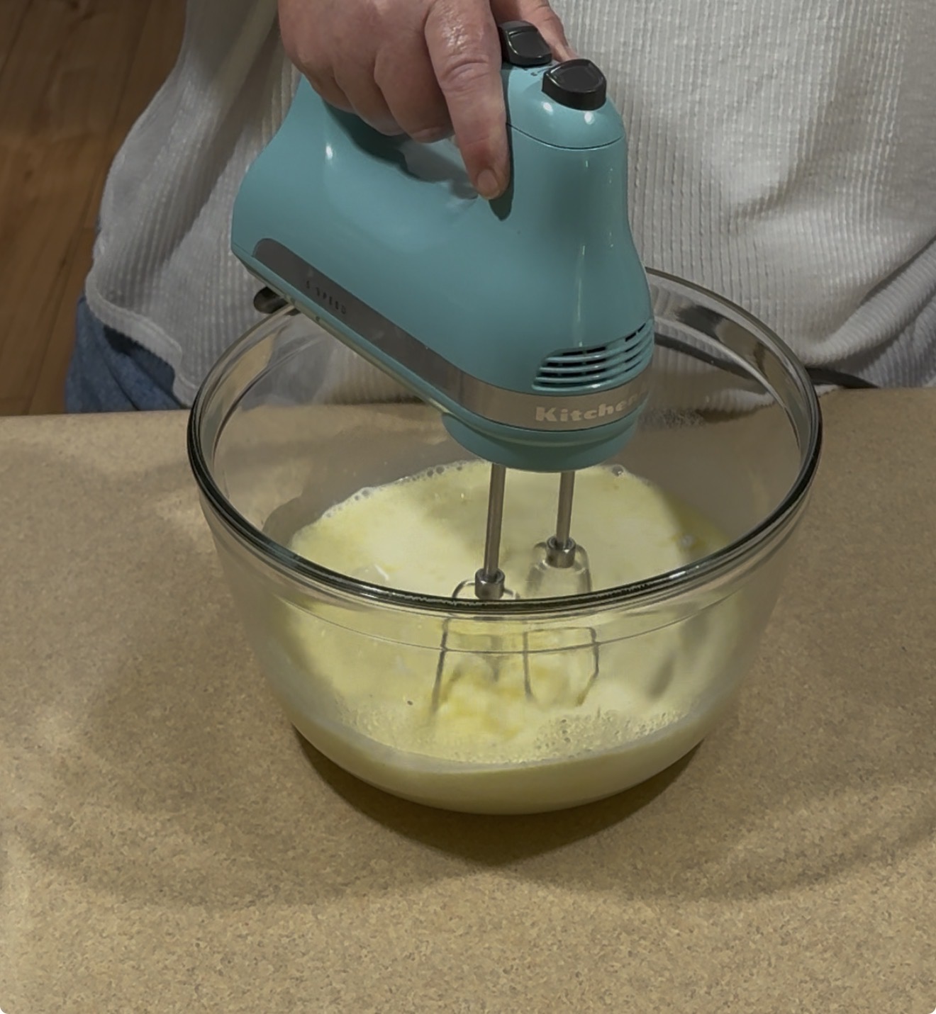 A person uses a blue electric hand mixer to beat eggs in a glass bowl on a kitchen counter.