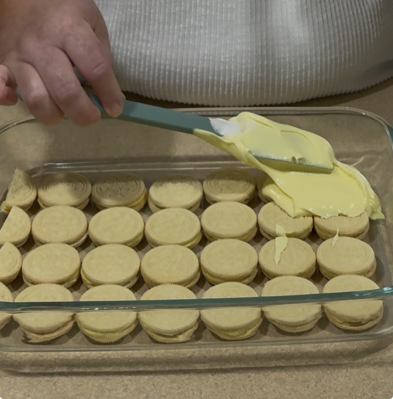 A person spreads yellow cream over a layer of vanilla sandwich cookies arranged in a glass baking dish.