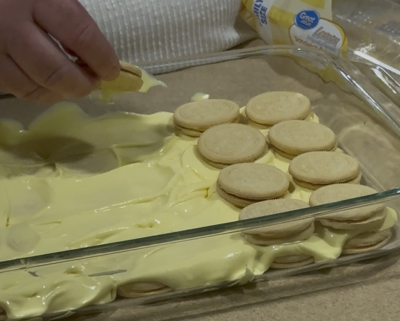 A hand places a vanilla sandwich cookie on top of a layer of cookies and yellow pudding in a glass baking dish, preparing a dessert. An open package of cookies is visible in the background.