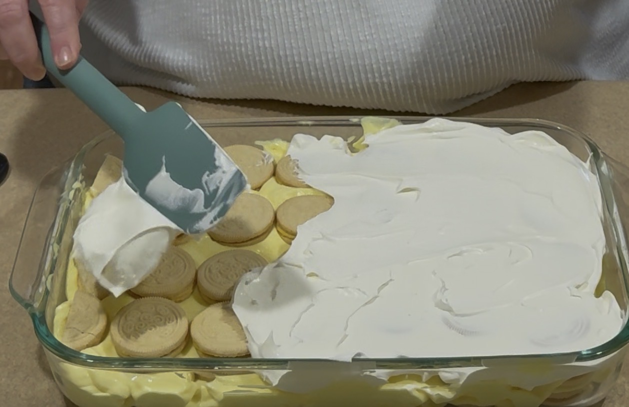 A person spreads whipped topping over a layer of yellow sandwich cookies and pudding in a glass baking dish using a spatula.