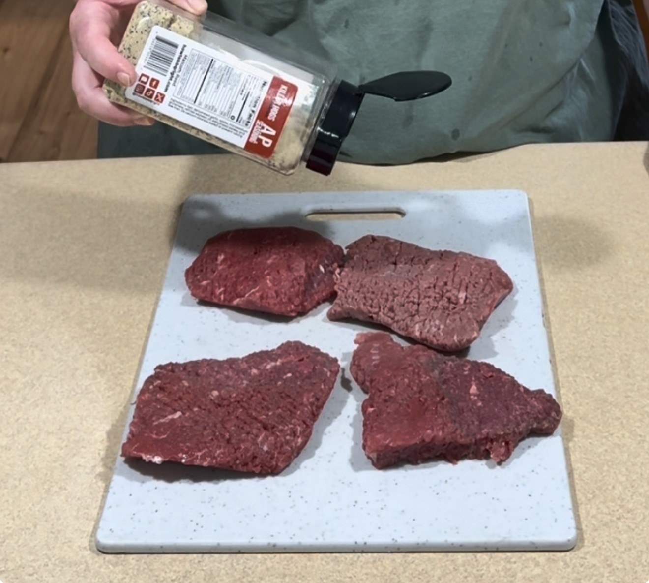 A person sprinkles seasoning from a container onto four raw beef steaks arranged on a white cutting board placed on a kitchen counter.