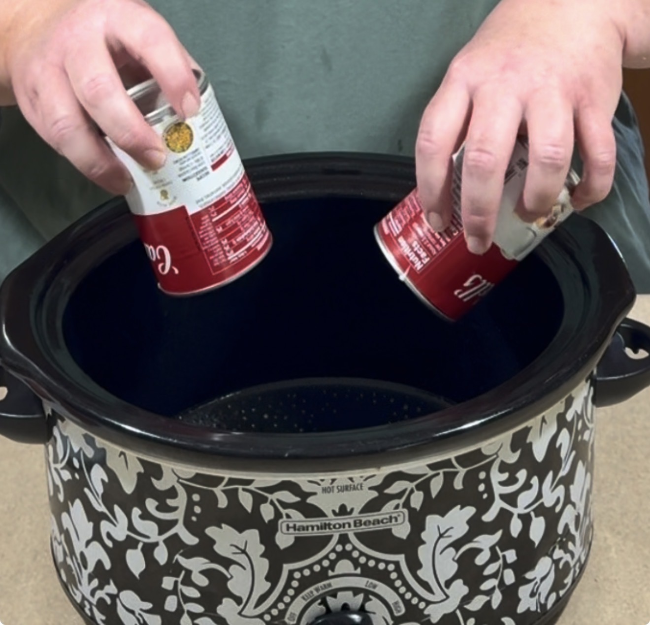 A person holding two cans of Campbell’s soup over an empty, black-and-white patterned Hamilton Beach slow cooker.