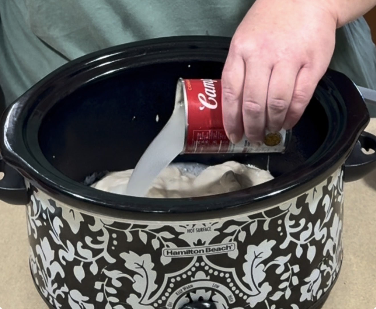 A person pours Campbell’s soup from a can into a black and white patterned Hamilton Beach slow cooker containing a creamy mixture.