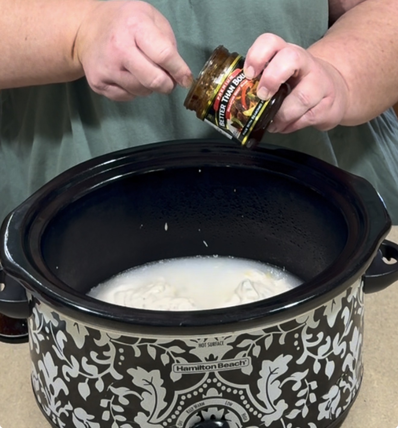 A person is adding a spoonful of seasoning from a jar into a black and white Hamilton Beach slow cooker filled with a creamy liquid mixture.
