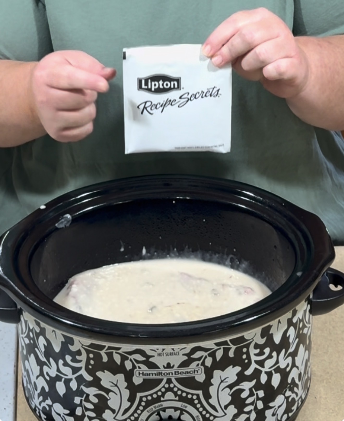 A person holds a Lipton Recipe Secrets packet above an open slow cooker containing a creamy mixture. The slow cooker is patterned and labeled Hamilton Beach.