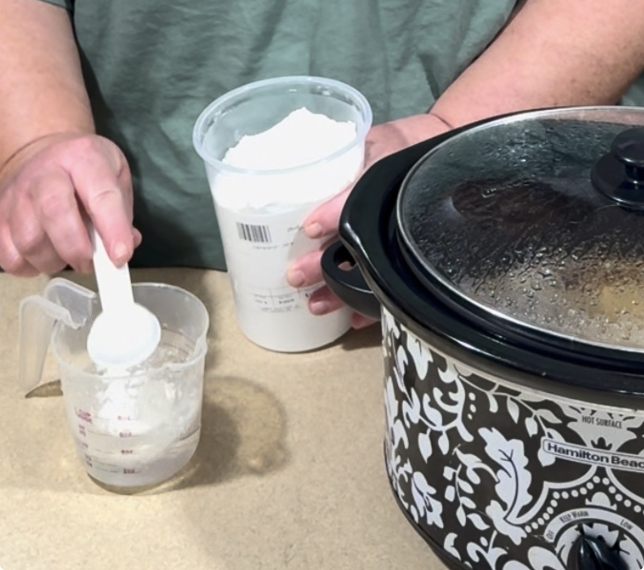 A person scoops white powder, likely flour or cornstarch, from a plastic container into a measuring cup, next to a patterned slow cooker on a countertop.