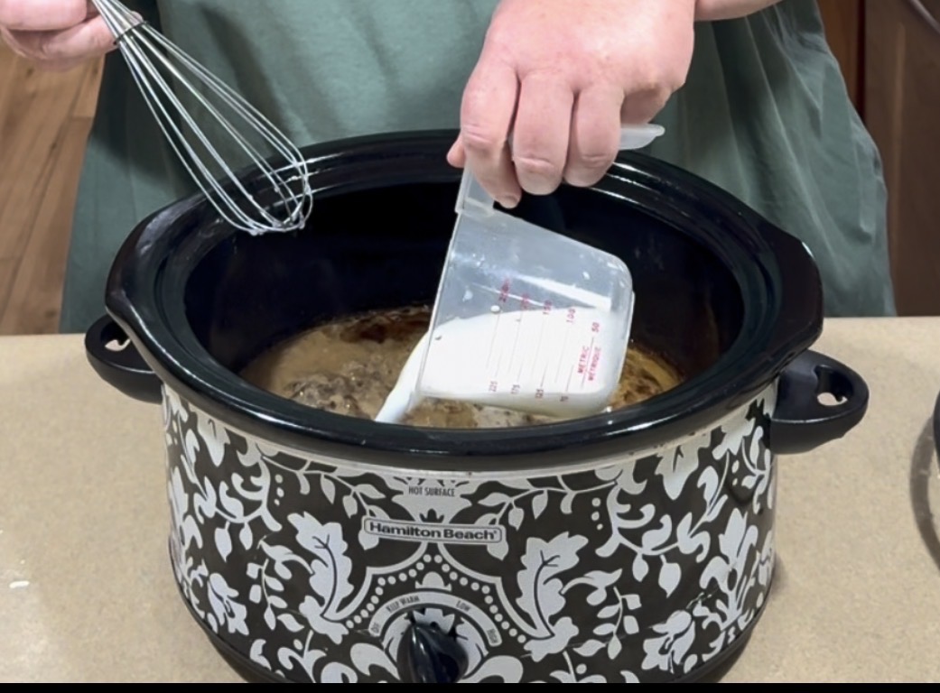 A person pours liquid from a measuring cup into a patterned slow cooker while holding a whisk over the pot. The slow cooker sits on a countertop.