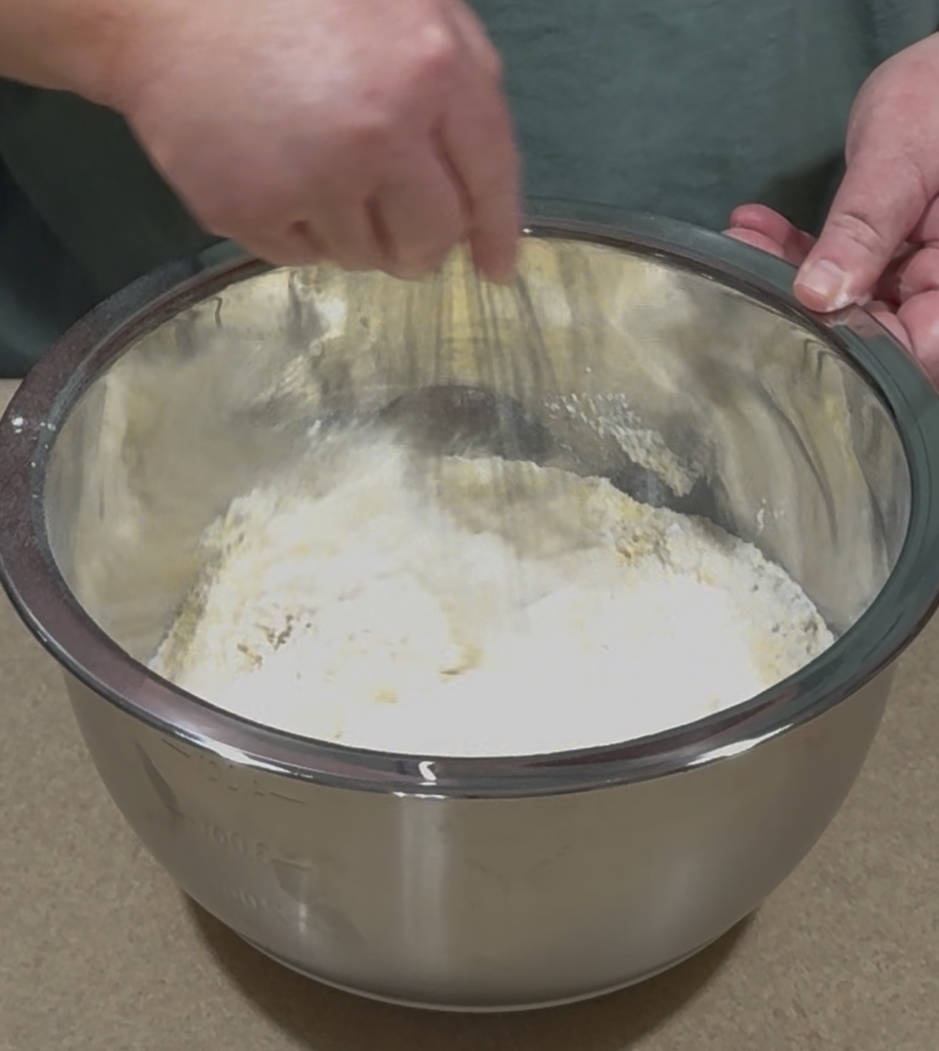 A person is whisking flour in a large stainless steel mixing bowl on a countertop.