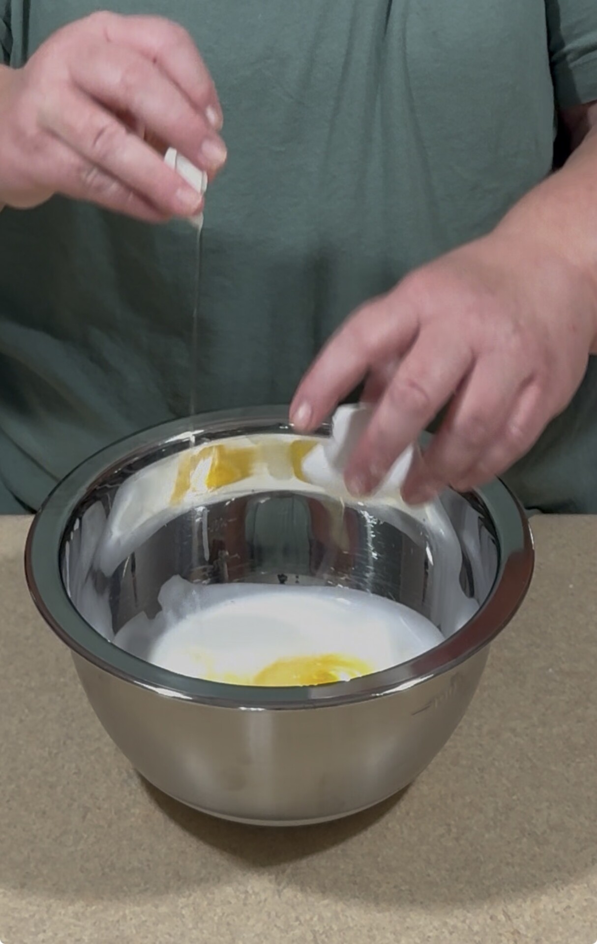 A person is cracking an egg into a large metal mixing bowl already containing sugar and eggs, preparing ingredients on a kitchen counter.