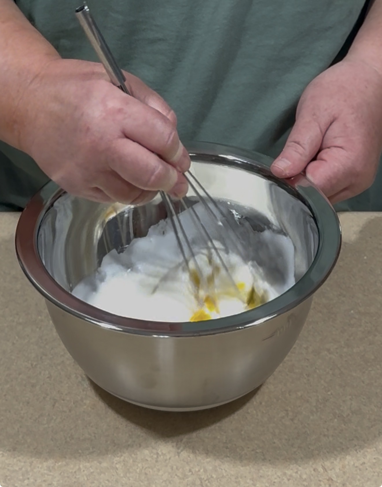 A person whisks egg yolk into a bowl of whipped egg whites using a metal whisk, with both hands holding the bowl and whisk over a countertop.