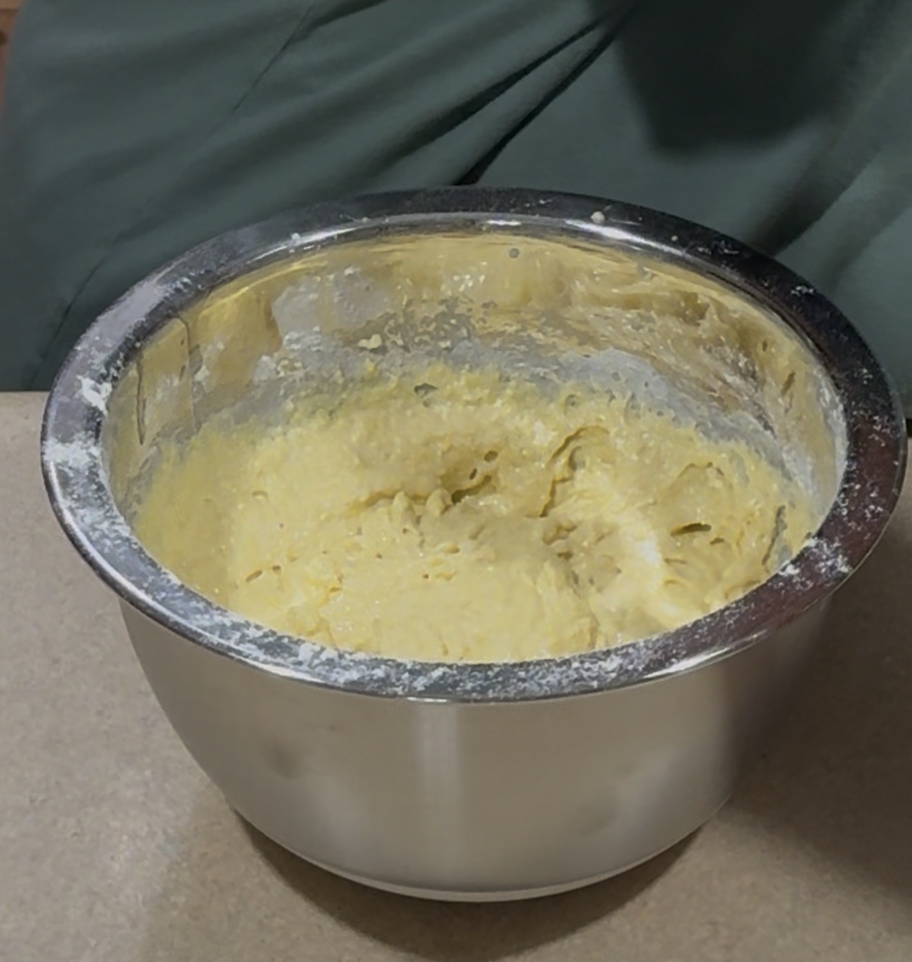 A large metal mixing bowl filled with thick, pale yellow batter sits on a kitchen counter, with some batter and flour visible on the rim.
